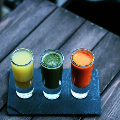 Zu sehen sind drei Shotgläser aus Glas. Sie sind gefüllt mit gelber, orangener und grüner Flüssigkeit. Der Hintergrund ist ein Holztisch. (Foto: Unsplash/Toa Heftiba)
