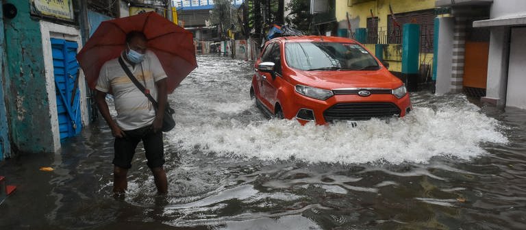 COP27: Ein Auto fährt durch eine überschwemmte Straße, in der ein Mann mit Regenschirm steht. In Kalkutta ist ein Starkregen niedergegangen, der mehrere Teile der Stadt überschwemmt hat. (Foto: dpa Bildfunk, picture alliance/dpa/ZUMA Wire | Debarchan Chatterjee)