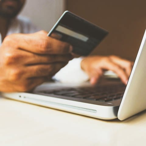Ein Mann hält eine Kreditkarte in der Hand, vor ihm ist ein Laptop geöffnet (Foto: Unsplash/ rupixxen)