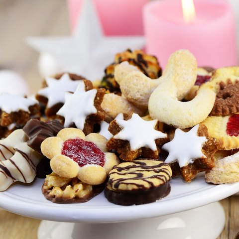 Verschiedene Plätzchen zu Weihnachten wie Vanillekipferl, Zimtsterne, Plätzchen mit Schokolade oder Marmelade liegen auf einer weißen Platte. Im Hintergrund  brennt eine pinke Kerze. (Foto: Adobe Stock/Printemps)