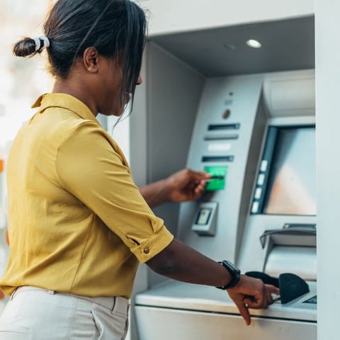 Dunkelhäutige Frau steht vor einem Bankautomaten. Mit ihrer linken Hand schiebt sie die Bankkarte in den Automaten, mit der rechten Hand gibt sie die PIN ein. (Foto: AdobeStock/Zamrznuti tonovi)