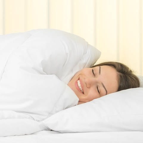 Eine Frau liegt im Bett. Ihr Kopf liegt auf einem Kissen. Ihr gesamter Körper ist von der Bettdecke umhüllt. (Foto: Adobe Stock, realstock1)