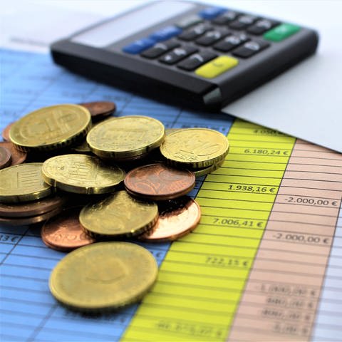 Auf einer Übersichtstabelle liegt Kleingeld, im Hintergrund ein Gerät für Online-Banking. (Foto: Colourbox, X28460681)