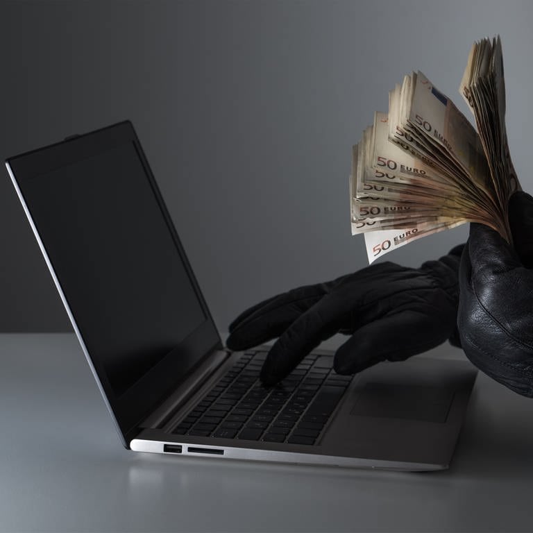 Person mit schwarzen Handschuhen hält Bündel Geldscheine vor einem Laptop. Ebay-Betrüger nutzen häufig dubiose Internetkonten. (Foto: Colourbox)