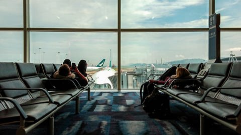 Zwei Reisende liegen auf Sitzmöglichkeiten am Flughafen und warten. Hinter der Scheibe ist ein Flugzeug mit Brücke zu sehen. (Foto: Unsplash / kirklai)