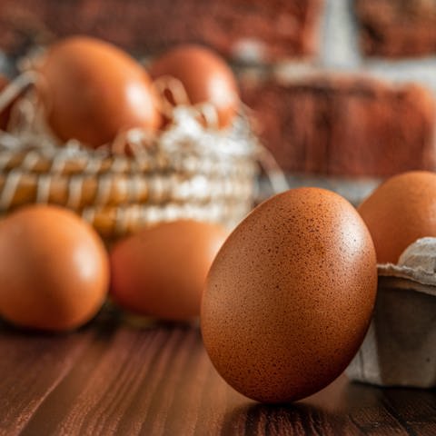Im Vordergrund lehnt ein braunes Ei an einem vollen Eierkarton. Im Hintergrund sind weitere Eier in einem Strohkorb zu sehen. (Foto: Adobe Stock)