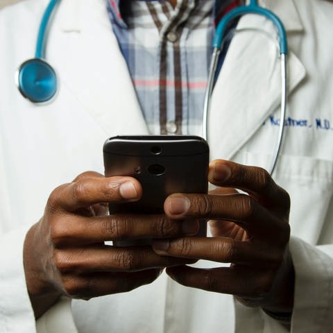 Ein Arzt in einem weißen Kittel hält ein schwarzes Smartphone in der Hand. Linksbündg steht der Beitagstitel: Termin buchen über Doctolib-Bald alternativlos? (Foto: Unsplash/National Cancer Institute)