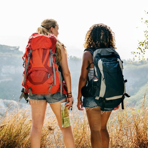 Zwei Frauen in Outdoorklamotten schauen in die Berge, mit einem günstigen roten Wanderrucksack und einem Trekkingrucksack (Foto: Adobe Stock)