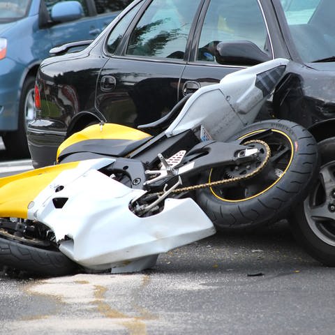 An einer Unfallstelle ist ein Motorrad mit einem Auto zusammengeprallt. Das Auto ist zerkratzt und Glas- und Motorradteile liegen auf der Straße. (Foto: Adobe Stock)