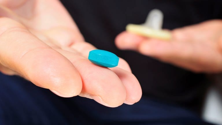 Auf einer Hand liegt eine blaue Viagra Pille, die gegen Erektionsprobleme helfen soll. Im HIntergrund ist ein Kondom zu sehen. (Foto: Colourbox/nito)