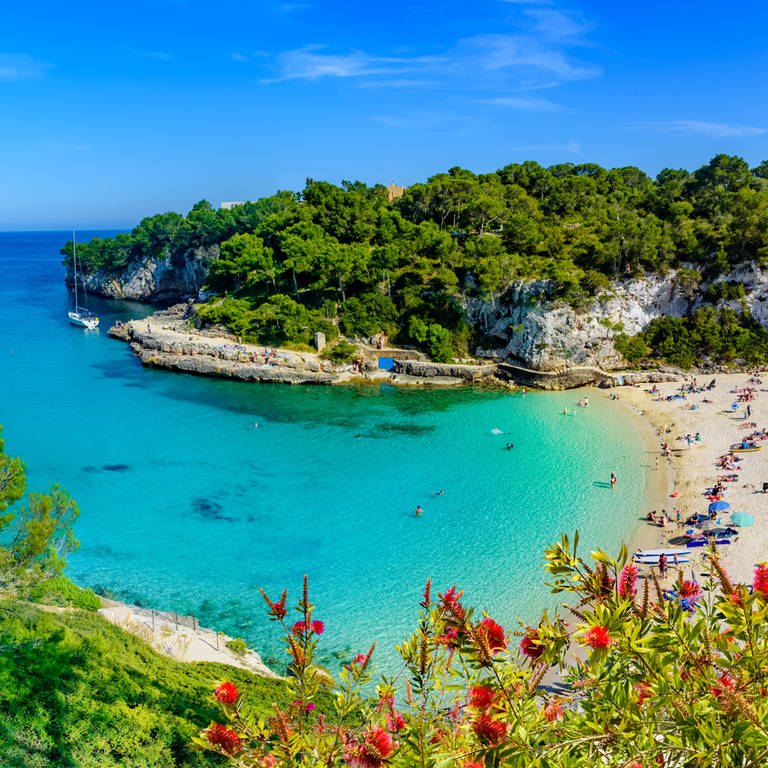 Das Bild zeigt den Ausblick über eine Bucht auf der Insel Mallorca. Das türkisne Wasser mündet ins Meer. Am STrand sind einige Menschen mit Sonnenschirmen zu erkennen. (Foto: Adobe Stock)