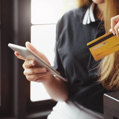 Frau mit langen dunkelblonden Haaren lehnt sich an eine Theke - in der einen Hand ihr Smartphone und in der anderen eine gelbe Kreditkarte. Smishing: Betrug mit Fake SMS und angeblichen Zoll-Gebühren (Foto: Adobe Stock, Adobe Stock/mojo_cp)