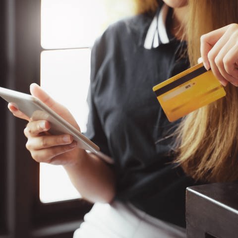 Frau mit langen dunkelblonden Haaren lehnt sich an eine Theke - in der einen Hand ihr Smartphone und in der anderen eine gelbe Kreditkarte. (Foto: Adobe Stock, Adobe Stock/mojo_cp)