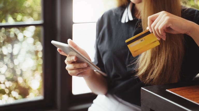 Frau mit langen dunkelblonden Haaren lehnt sich an eine Theke - in der einen Hand ihr Smartphone und in der anderen eine gelbe Kreditkarte. (Foto: Adobe Stock, Adobe Stock/mojo_cp)