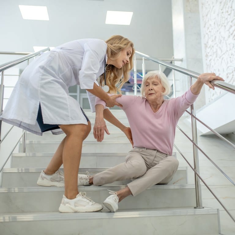 Ältere Dame sitzt auf Treppe und wird von junger Frau in weißem Kittel gestützt. Lohnen sich spezielle Senioren-Unfallversicherungen? (Foto: Adobe Stock)