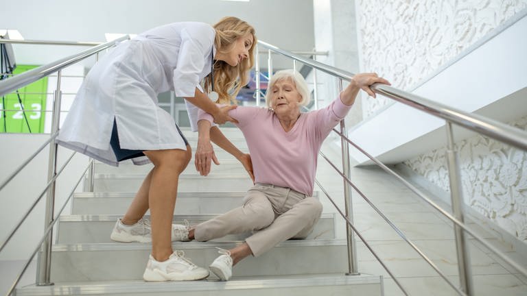 Ältere Dame sitzt auf Treppe und wird von junger Frau in weißem Kittel gestützt. Lohnen sich spezielle Senioren-Unfallversicherungen? (Foto: Adobe Stock)