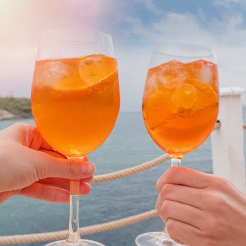Zwei Hände halten jeweils ein Weinglas mit dem Kult-Cocktail Aperol Spritz. Es ist nicht zu erkennen, dass es sich um eine günstigere Aperol Alternative handelt. Im Hintergrund ist das Meer zu sehen. (Foto: Adobe Stock)
