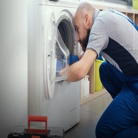 Handwerker mit Glatze und Bard trägt einen Blaumann und sitzt auf dem Boden rechts im Bild. Er repariert eine Waschmaschine (links im Bild). (Foto: Adobe Stock, Adobe Stock/StockPhotoPro)