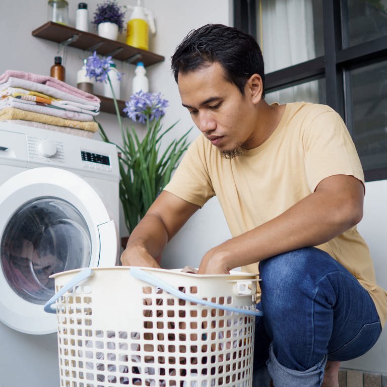 Junger Mann befüllt eine Waschmaschine mit Wäsche. Was sagt die Füllmenge bei der Waschmaschine aus? Welche ist wirklich effizient? (Foto: Adobe Stock)
