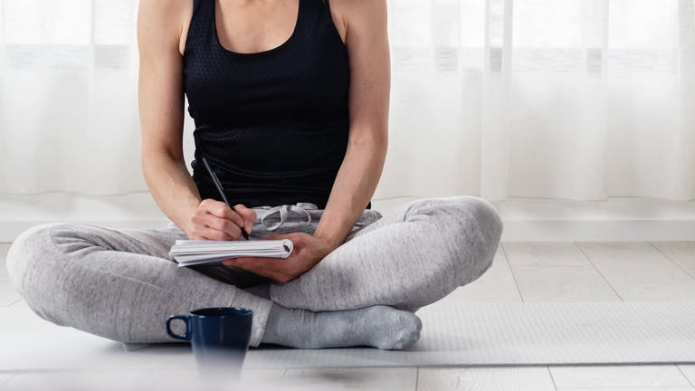 Frau sitzt auf einer Yoga-Matte, schreibt etwas auf
