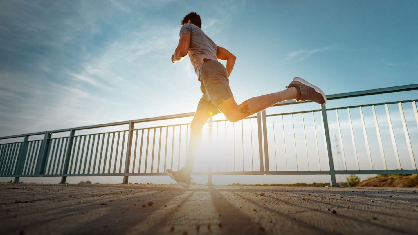 Mann joggt in Sportkleidung auf einer Brücke bei Sonnenschein. Warum ihr jetzt mit Sport anfangen solltet (Foto: Adobe Stock, bernardbodo)