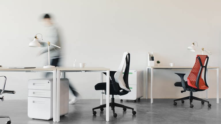 Zwei Bürotische samt Bildschirme und Stühle vor weißer Wand und eine verschwommene, laufende Person im Hintergrund  (Foto: Unsplash/Laura Davidson)