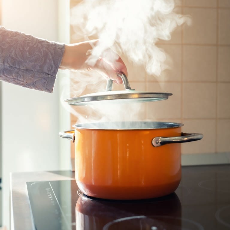 Energie- und Stromsparen in der Küche: Eine Hand setzt einen Deckel auf einen Kochtopf mit dampfendem Inhalt.
