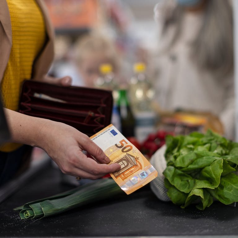 Person zahlt mit einem 50-Euro-Schein an der Kasser eines Supermarkts. Auf dem Kassenband liegt ein Salat und eine Lauchzwiebel, sowie weitere Produkte in der Unschräfe im Hintergrund.