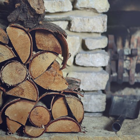 Auf dem Bild ist aufegstapeltes Brennholz vor einem Kamin.Im Hintergrund erkennt man eine Schaufel und eine Gabel für den Kamin und für das Feuer