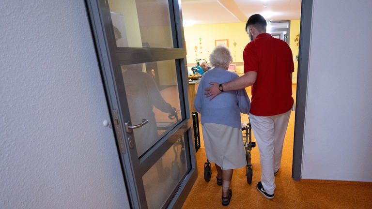 Eine ältere Dame mit Rollator und ein Pfleger gehen gemeinsam durch eine Tür