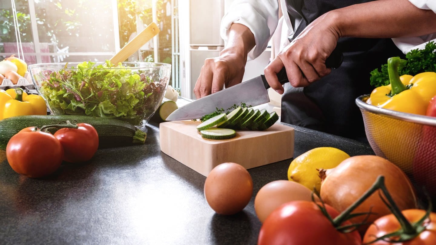 Person schneidet Gemüse in der Küche. Manche schwören auf Rohkost, andere vertragen Gemüse nur gegart. Welche Zubereitungsart die gesündeste ist und die Vitamine erhält – ist bei jedem Gemüse anders. (Foto: Adobe Stock)