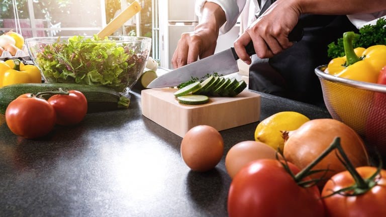 Person schneidet Gemüse in der Küche. Manche schwören auf Rohkost, andere vertragen Gemüse nur gegart. Welche Zubereitungsart die gesündeste ist und die Vitamine erhält – ist bei jedem Gemüse anders. (Foto: Adobe Stock)