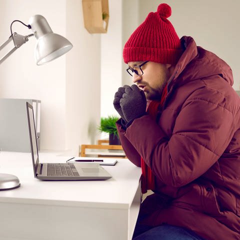 Ein Mann trägt eine rote Wollmütze auf seinem Kopf. Außerdem trägt er Handschuhe und eine Winterjacke. Er sitzt auf einem Stuhl, vor einem Schreibtisch, auf dem ein Laptop steht. Seine Hände hält er sich zum wärem vor den Mund.