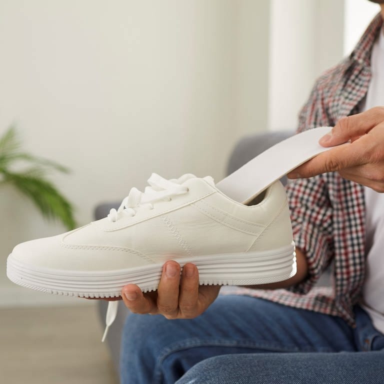 Ein Mann kniet auf dem Boden. Er hält einen weißen Sneaker in seiner rechten Hand. Mit seiner linken Hand schiebt er eine Einlegesohle in den Schuh.