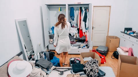 Eine Frau mit langen braunen Haaren ist von hinten zu sehen, wie sie vor einem chaotischen Kleiderschrank steht, überall liegen Taschen und Kleidungsstücke herum. Zuschaueraufruf: Marktcheck sucht Protagonisten