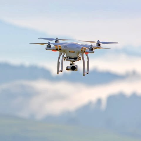 Eine weiße Drohne mit vier Rotoren und einer angebauten Kamera fliegt in der rechten oberen Ecke des Bildes vor einer unscharfen Landschaft im Hintergrund