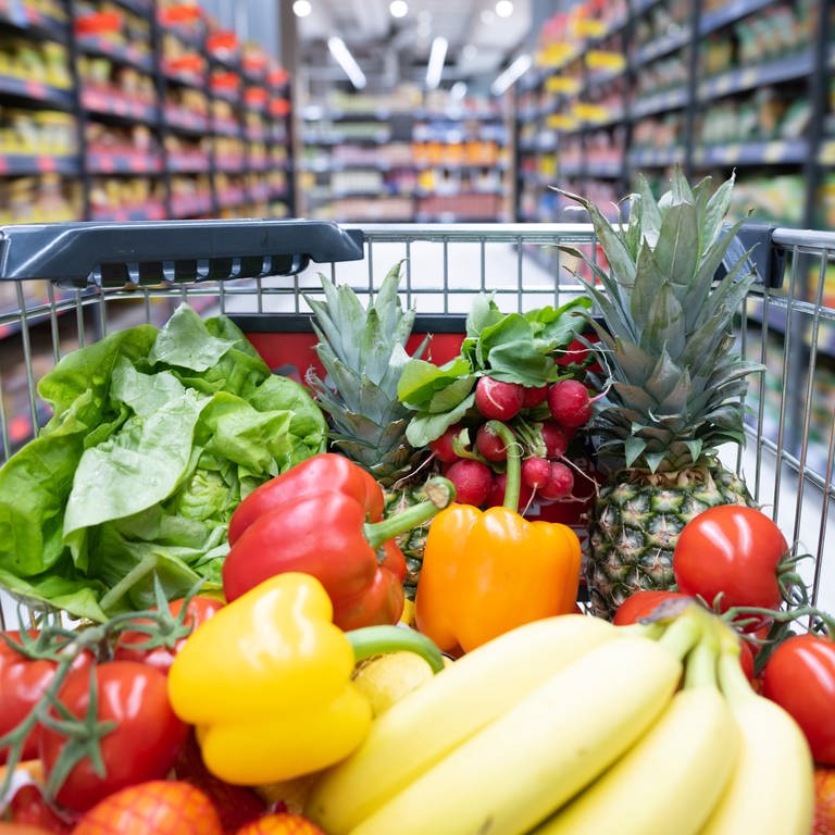 Ein Einkaufskorb mit Obst und Gemüse steht in einem Supermarkt. 