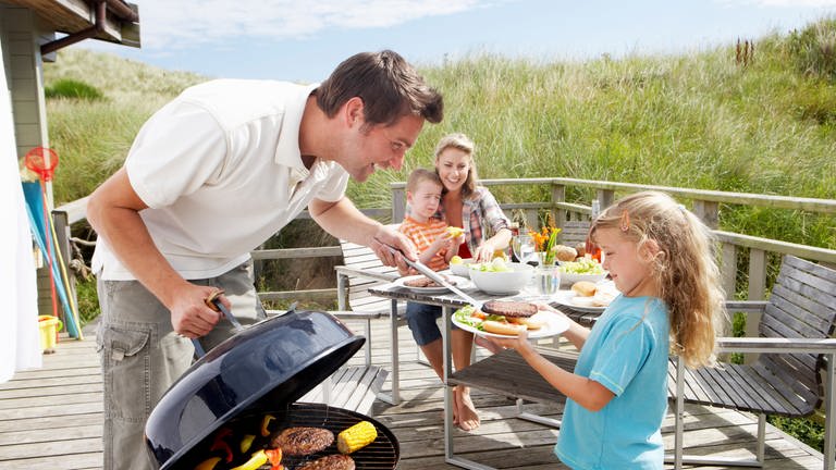 Eine Familie grillt. Der Mann gibt seiner kleinen Tochter ein Steak. Auf dem Grill liegen Grillfleisch und Mais. Frau und Sohn sitzen am Tisch im Hintergrund.