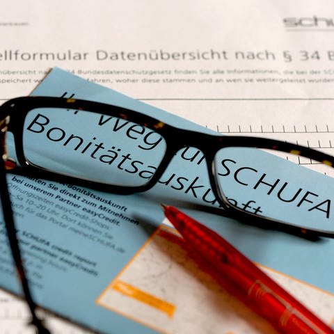 Auf einem Antrag für die Datenkopie der Auskunftei Schufa liegt ein blauer Flyer zum Thema Schufa-Bonitätsauskunft, eine Brille und ein roter Kugelschreiber.