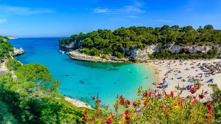 Das Bild zeigt den Ausblick über eine Bucht auf der Insel Mallorca. Das türkisne Wasser mündet ins Meer. Am STrand sind einige Menschen mit Sonnenschirmen zu erkennen.