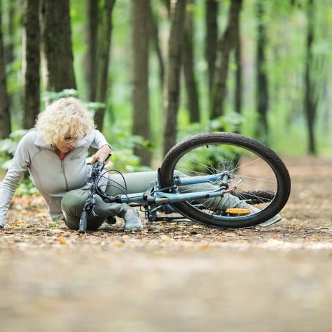 Eine Frau stürzt auf einem Waldweg mit ihrem Fahrrad und versucht sich aufzustemmen wähernd ihr Bein unter dem blauen Fahrrad liegt. Was tun, wenn die Senioren-Unfallversicherung nicht zahlt?