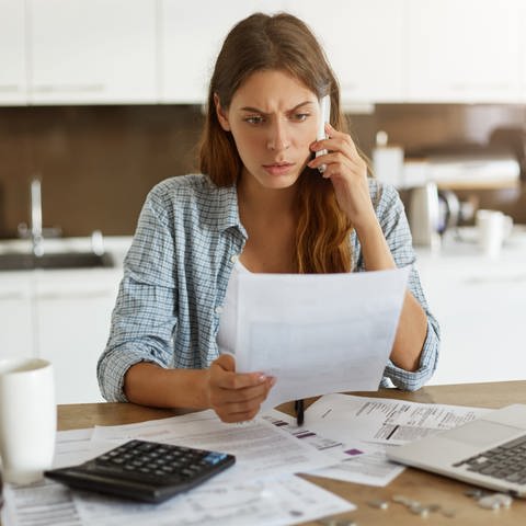 Eine besorgte junge Frau sitzt an einem Schreibtisch voller Rechnungen und hält ein Blatt Papier in der Hand während sie telefoniert.