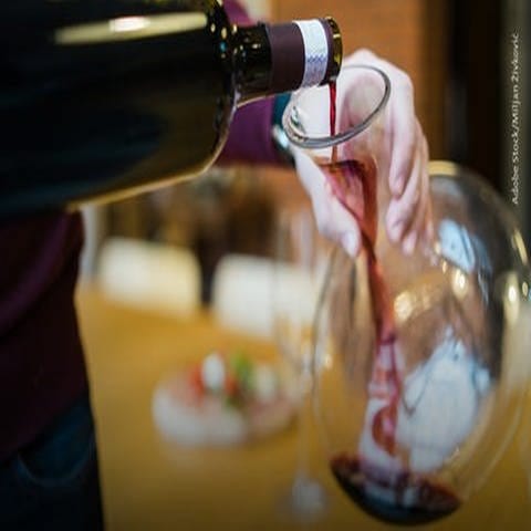 Links im Bild ist eine Rotweinflasche zu sehen, deren Inhalt in einen Weindekanter (rechts im Bild) gegossen wird.