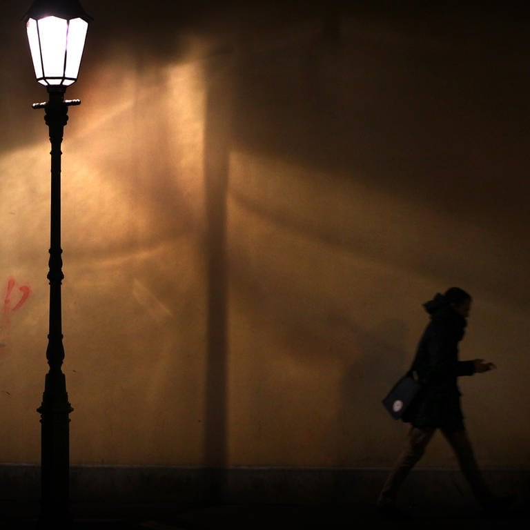 Eine junge Frau im Licht einer Straßenlaterne auf einem Bürgerseig: Viele Menschen haben Angst davor, nachts im Dunkeln nach Hause zu gehen. Das Heimwegtelefon, Apps oder Frauentaxis können helfen. 