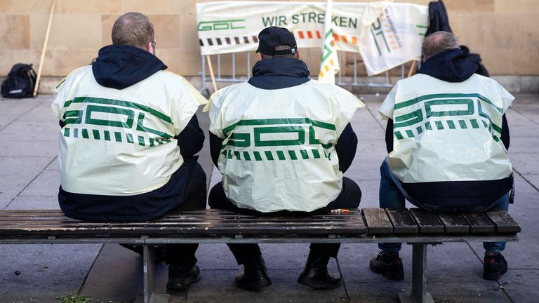 Teilnehmer eines Warnstreiks der Gewerkschaft Deutscher Lokomotivführer (GDL) sitzen vor dem Hauptbahnhof auf einer Bank.