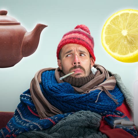 Bärtiger Mann sitzt mit Fieberthermometer im Bund, Mütze und in Decken gehüllt da und blickt wehleidig nach oben. Welche Hausmittel helfen wirklich gegen Erkältung?