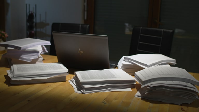 Mehrere Stapel Papier liegen um einen Laptop herum auf einem Tisch.