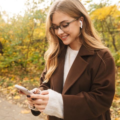Frau mit blonden, langen Haaren und einer Brille schaut auf ihr Smartphone trägt dabei Airpods
