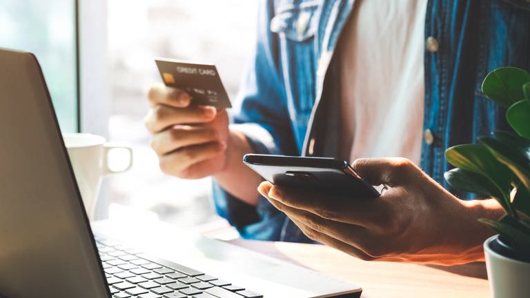 Person hält eine Bankkarte und ein Smartphone in der Hand und sitzt vor einem Laptop. Phishing: Wenn der Online-Shop Daten abgreift (Foto: Adobe Stock, Thapana_Studio)