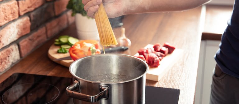 Spaghetti werden in einen Topf heißes Wasser gegeben. Im Hintergrund sind weitere Zutaten für ein Nudelgericht zu sehen. Tipps zum Nudelkochen erfahren Sie bei uns. (Foto: Colourbox, COLOURBOX48225180)